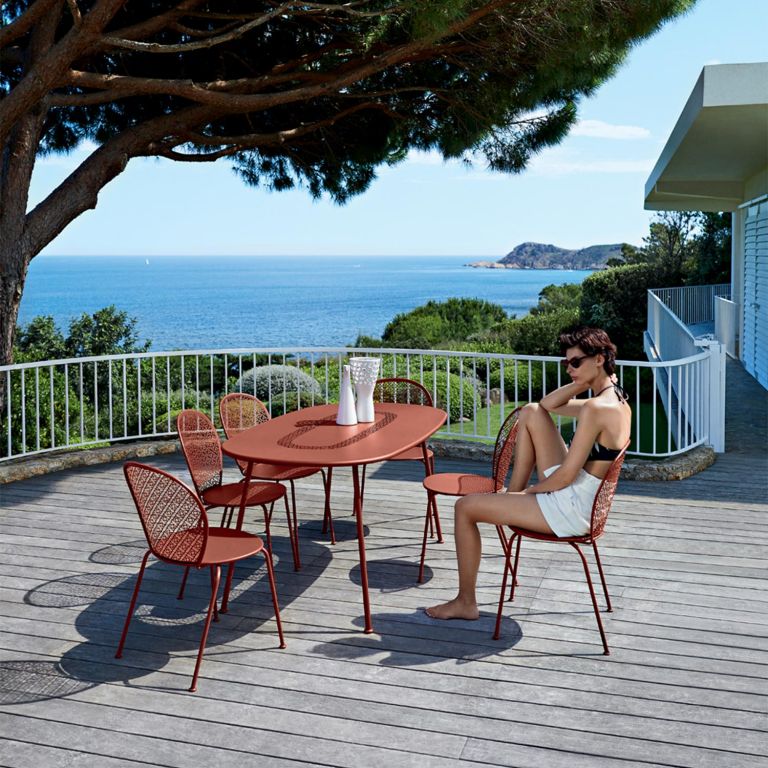 Woman sits at red ochre Fermob Lorette oval outdoor table and chairs on a deck terrace overlooking sea